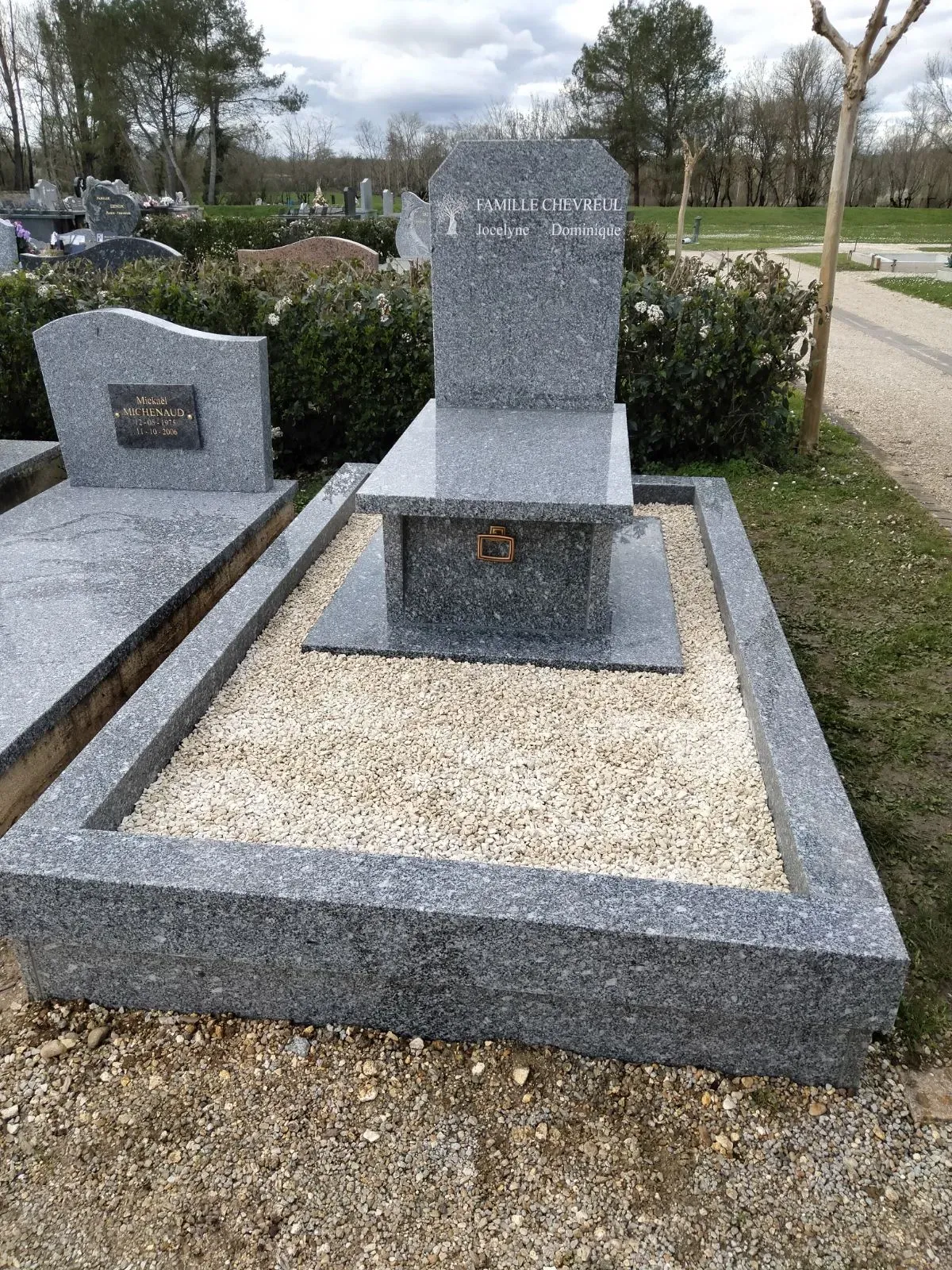 Monument funéraire en granit gris gravé entouré de graviers clairs dans un cimetière.